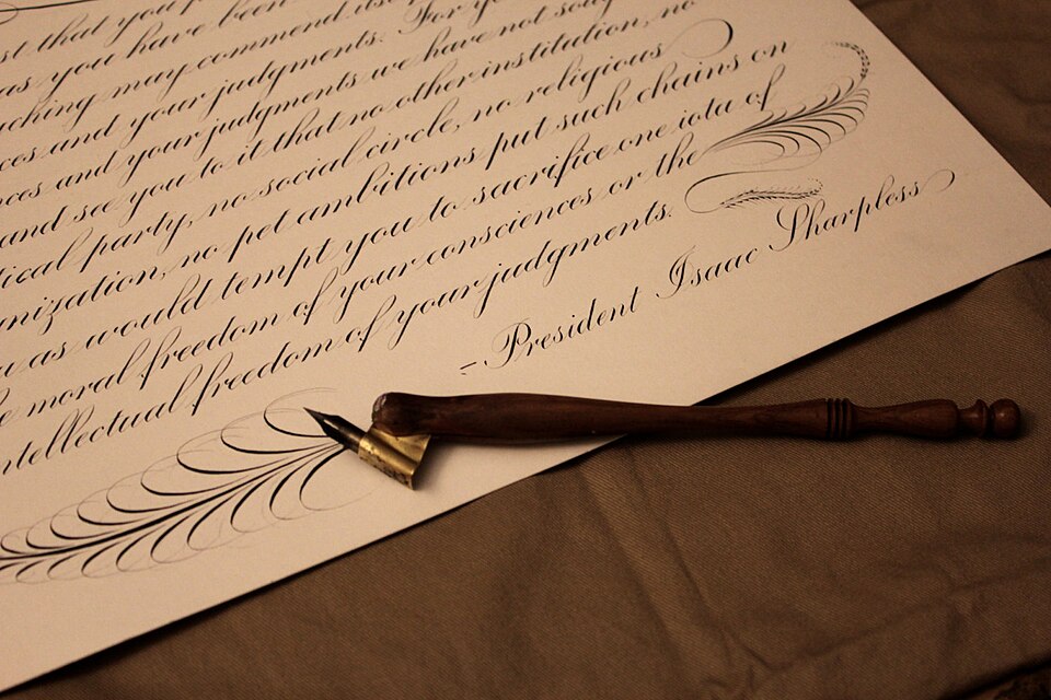 A sample of copperplate script with offhand flourishing. The pen-holder in the photo (an oblique pen-holder) is commonly used by copperplate calligraphers. The quotation is by Isaac Sharpless, president of Haverford College, from a speech given in 1888.